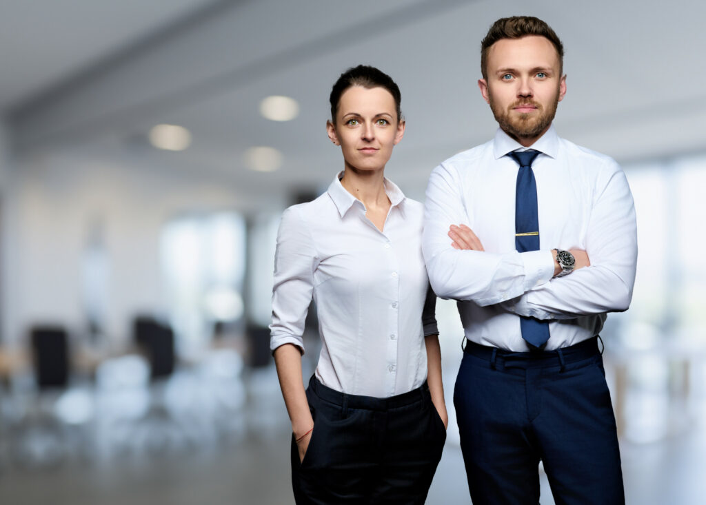 Zwei Personen in Business-Kleidung stehen selbstbewusst in einem hellen, modernen Büro. Die Person links, möglicherweise ein Anwalt, hat die Hände in den Taschen und trägt ein helles Hemd und eine dunkle Hose. Die Person rechts verschränkt die Arme mit Krawatte und Uhr. Der Hintergrund ist leicht unscharf.