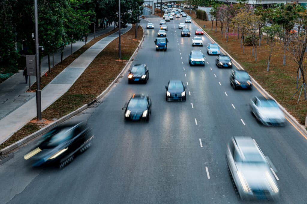 Eine vielbefahrene, mehrspurige Straße mit zahlreichen Autos in beiden Richtungen, deren Bewegungen wie die schnellen Entscheidungen eines Anwalts verschwimmen. Bäume und ein Bürgersteig säumen die Straße. Der Himmel ist bedeckt, und Straßenlaternen wachen über die belebte Straße.