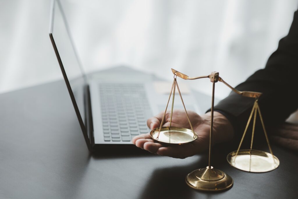 Eine Person hält eine kleine goldene Waage neben einem offenen Laptop auf einem Schreibtisch. Sie symbolisiert die Verbindung von Recht und Technologie, die den modernen Anwalt ausmacht. Der Hintergrund ist sanft beleuchtet und schafft eine professionelle und zugleich besinnliche Atmosphäre.